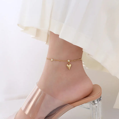 Bracelet de cheville en acier inoxydable doré pendentif cœur et zircons cubiques autour d'une cheville de femme avec une robe blanche et des sandales Kaosix