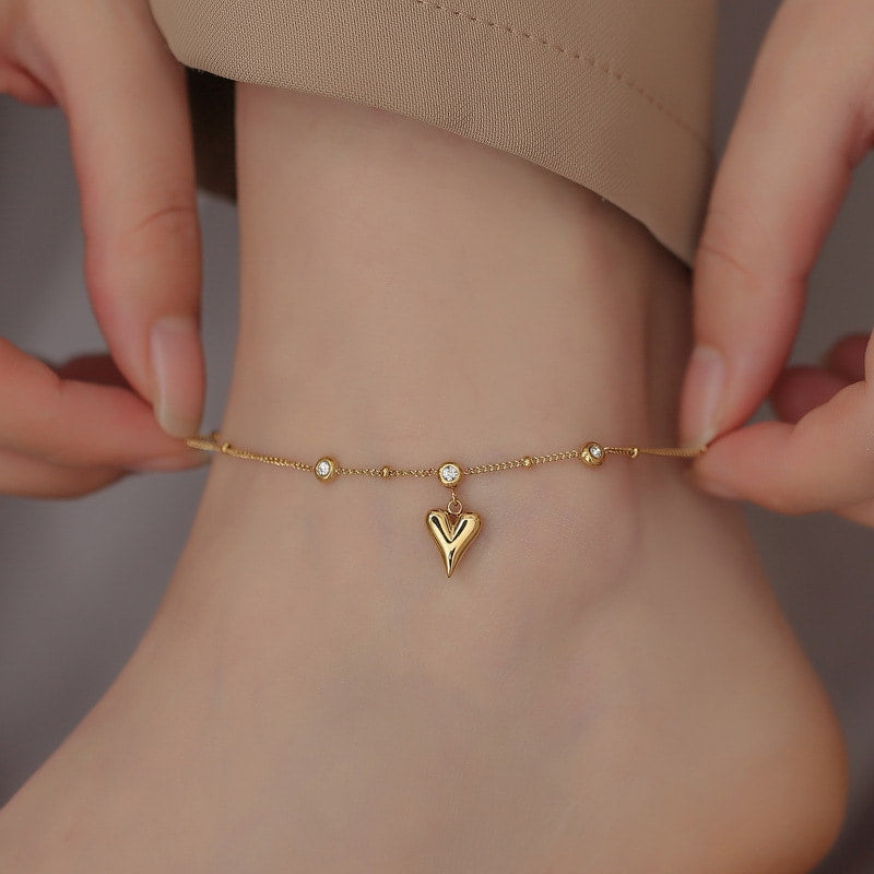 Bracelet de cheville en acier inoxydable doré pendentif cœur et zircons cubiques autour d'une cheville de femme qui porte un pantalon beige Kaosix