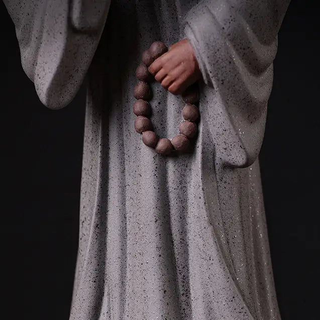 Statue Moine Bouddhiste Humilité et Respect Debout Blanc sur fond noir vue en gros plan de la main gauche tenant un mala de perles marron Kaosix