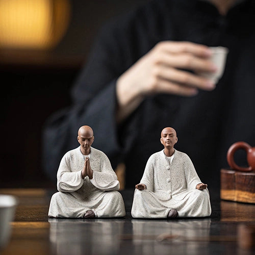 Deux Statues Moines Bouddhistes Prière et Méditation posés sur une table en bois avec un homme qui boit du thé en arrière plan kaosix