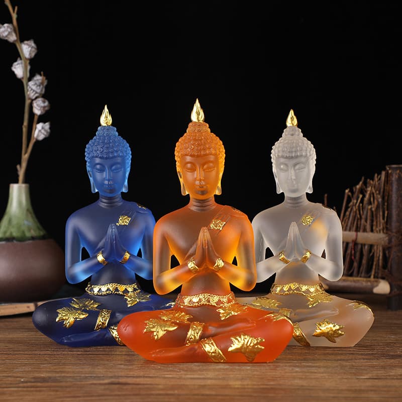 Trois Statuettes de Bouddha en résine translucide bleu orange blanc assises de face sur une table en bois et un fond noir Kaosix