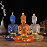 Trois Statuettes de Bouddha en résine translucide bleu orange blanc assises de face sur une table en bois et un fond noir Kaosix