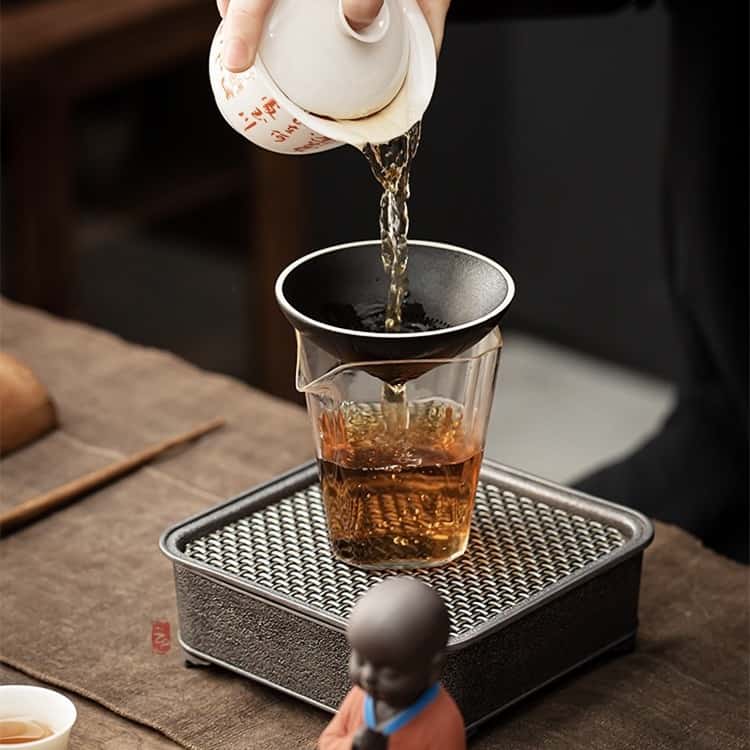 Une main qui verse du thé depuis une théière dans un verre avec la chapeau chinois qui sert de filtre pour le thé Kaosix