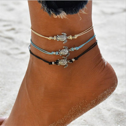 3 bracelets de cheville avec amulette tortue de mer en métal argenté et cordon bleu blanc ou noir autour de la cheville d'une femme sur la plage Kaosix