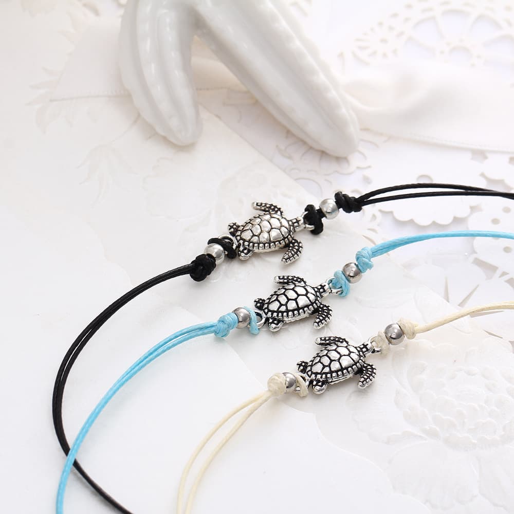 3 bracelets de cheville avec amulette tortue de mer en métal argenté et cordon bleu blanc ou noir sur une nappe blanche et gros plan sur les amulettes tortues Kaosix