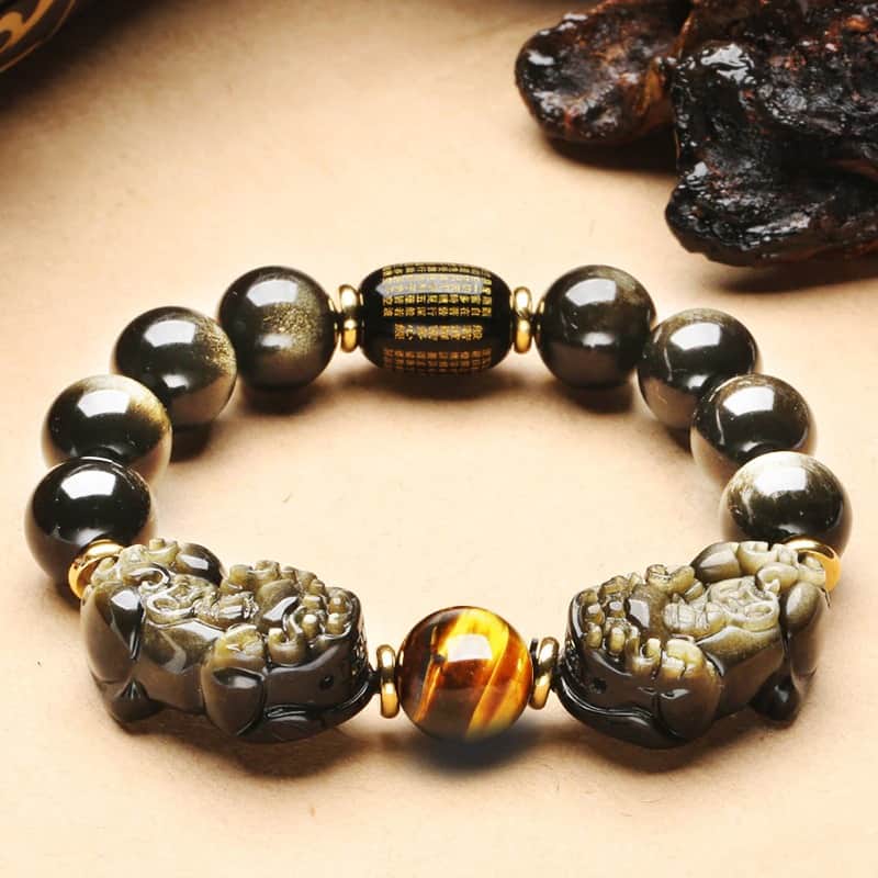 Bracelet Double Pi Xiu (Pi Yao) Obsidienne Dorée Œil de Tigre posé sur un sol beigeavec un rocher noir en arrière plan Kaosix