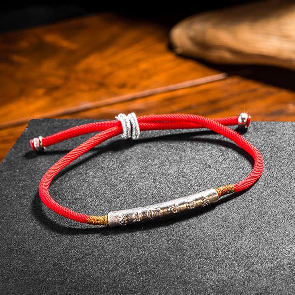 Bracelet Tibétain Rouge Chance et Mani Mantra argenté sur un socle gris foncé et posé sur une table en bois Kaosix