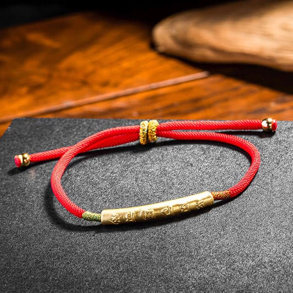Bracelet Tibétain Rouge Chance et Mani Mantra doré sur un socle gris foncé et posé sur une table en bois Kaosix