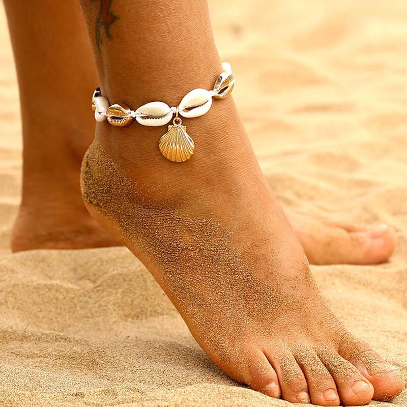 Bracelet de cheville en coquillages cauris avec amulette coquille Saint-Jacques couleur or autour de la cheville d'une femme sur la plage dans le sable Kaosix