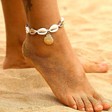 Bracelet de cheville en coquillages cauris avec amulette coquille Saint-Jacques couleur or autour de la cheville d'une femme sur la plage dans le sable Kaosix