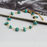 Bracelet de cheville en éclats de pierre turquoise et chaînette dorée posé sur une table en bois Kaosix