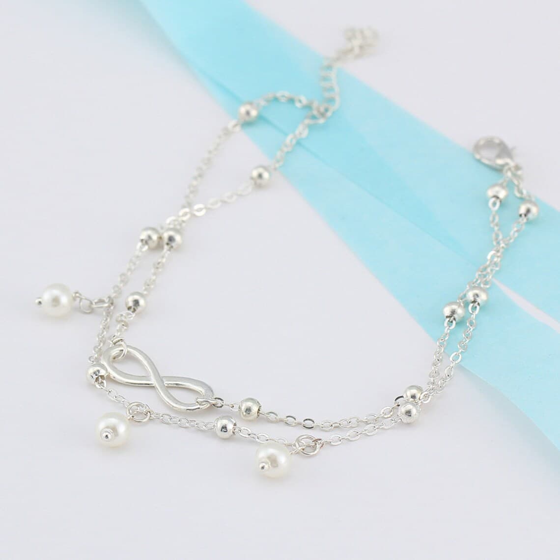 Bracelet de cheville infini et perles chainette argentée sur une nappe grise Kaosix