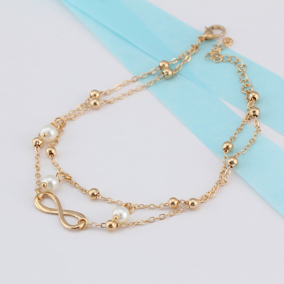 Bracelet de cheville infini et perles chainette dorée sur une nappe grise Kaosix