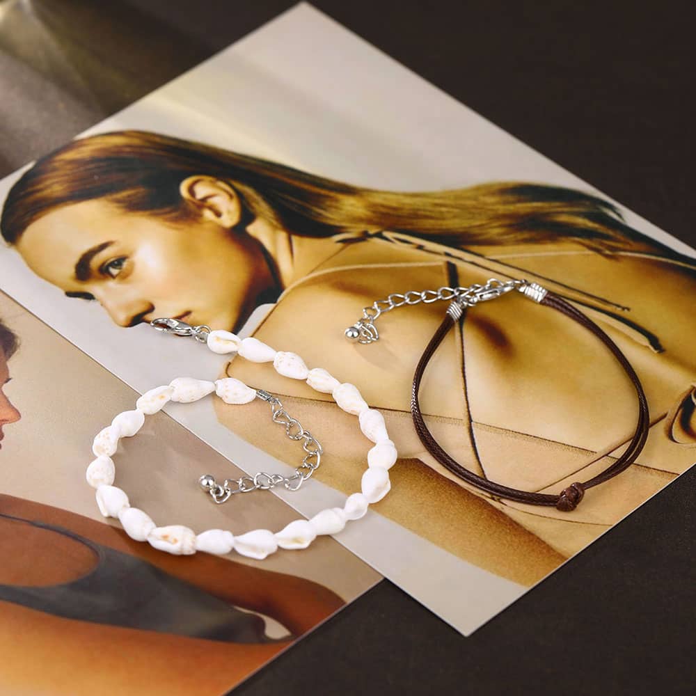 Deux braceleta de cheville en coquillages buccin et cordon de cuir posé sur un magazine ouvert Kaosix