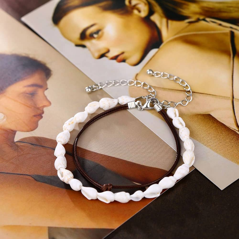 Double bracelet de cheville en coquillages buccin et cordon de cuir posé sur un magazine ouvert Kaosix