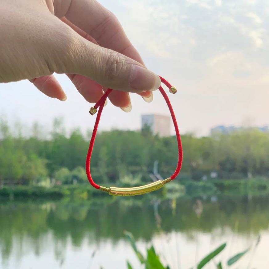 Homme tenant le Bracelet Tibétain Rouge Chance et Mani Mantra doré dans sa main avec un lac en fond Kaosix