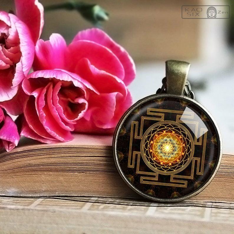 Pendentif Sri Yantra en verre à géométrie sacrée posé contre un livre ouvert et avec une rose en arrière-plan kaosix