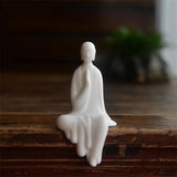 Statue moine bouddhiste zen céramique blanche pose Abhaya mudra assis sur banc en bois avec fond noir Kaosix